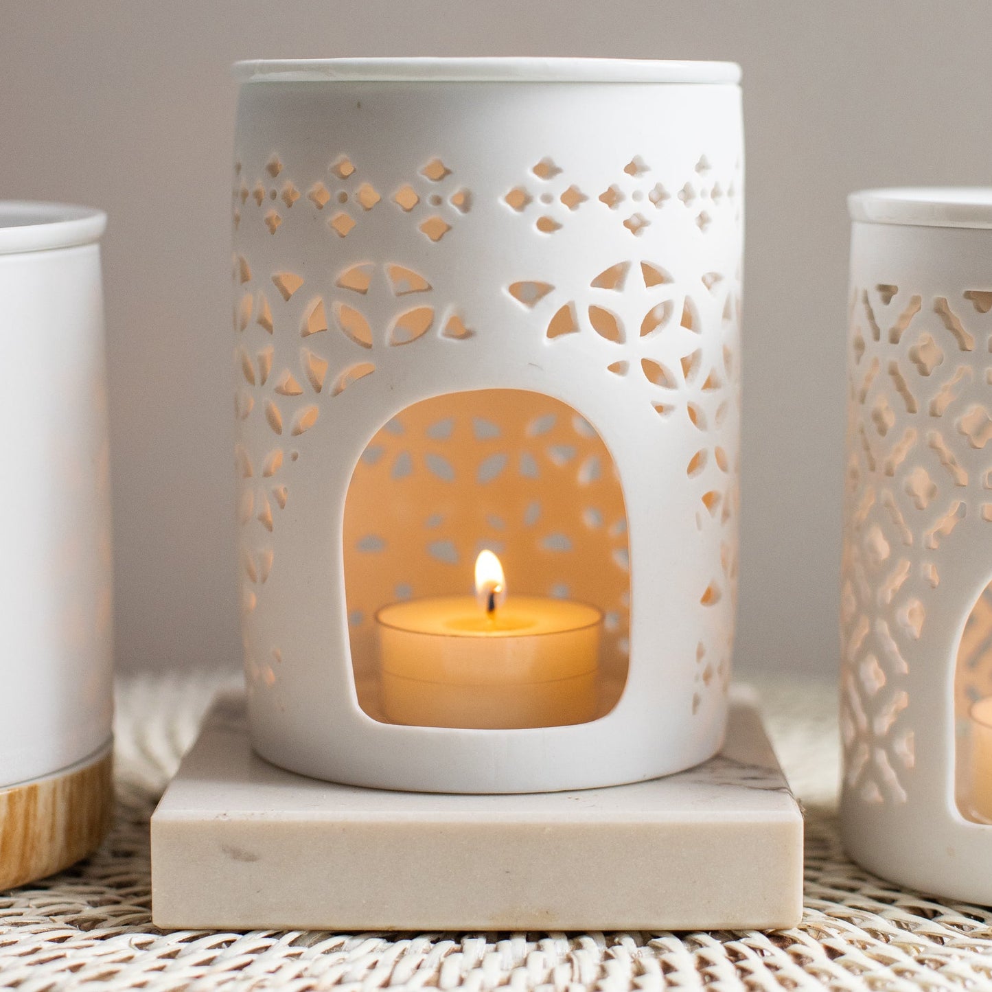 White ceramic lantern with cut-out patterns holding a lit candle on a textured surface.