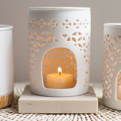 White ceramic lantern with cut-out patterns holding a lit candle on a textured surface.