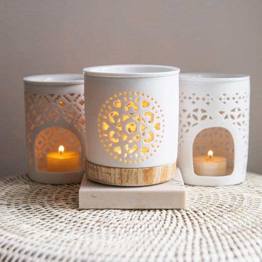 Three white ceramic lanterns with decorative cutouts, each containing a lit candle, on a textured surface.