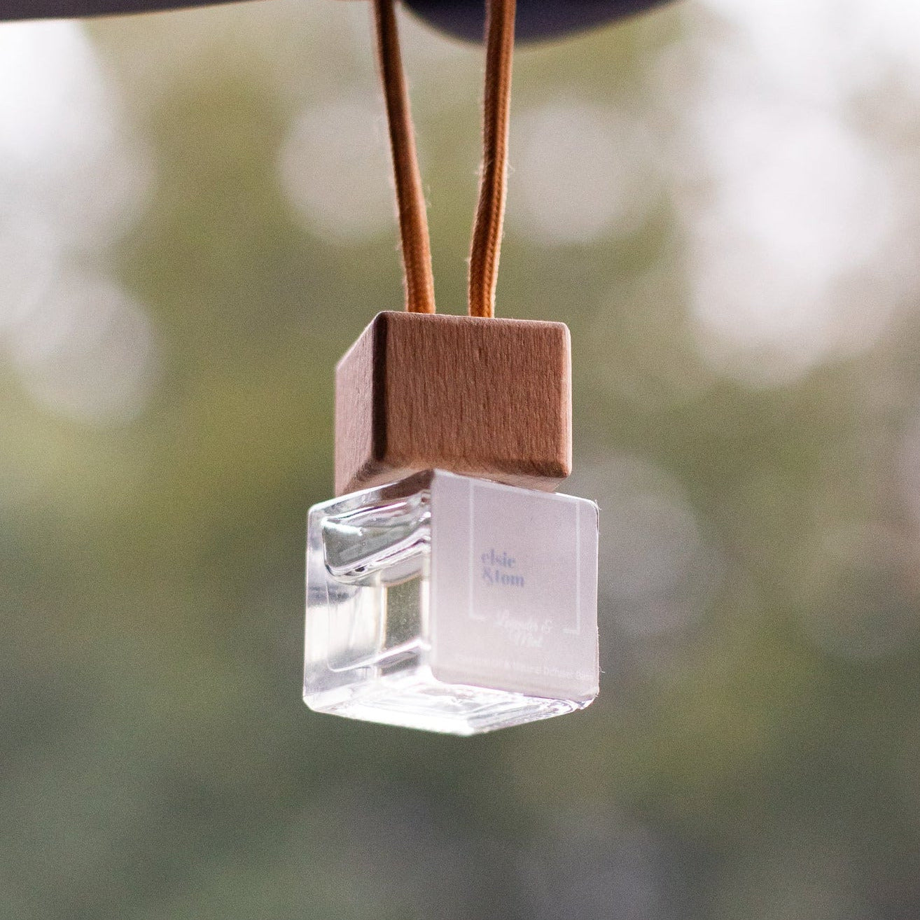 Natural Aromatherapy Scented Car Diffuser air freshener hanging from a car's rear view mirror with a blurred green background