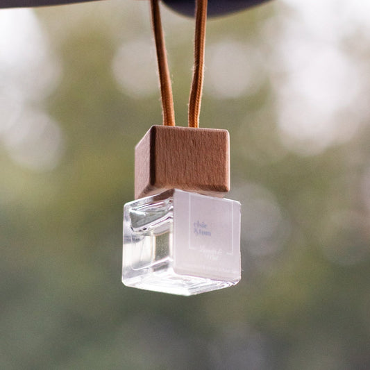 Natural Aromatherapy Scented Car Diffuser air freshener hanging from a car's rear view mirror with a blurred green background